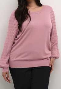 Pink sweater with a smooth body and textured, ribbed sleeves. Rounded neckline and fitted cuffs, paired with black pants.