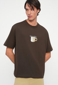 Καφέ βαμβακερό T-shirt με κοντά μανίκια, που διαθέτει ένα κεντημένο σχέδιο με καφέ φλιτζάνι και το κείμενο "Café du Choux" σε ανοιχτό μπλε από πάνω.