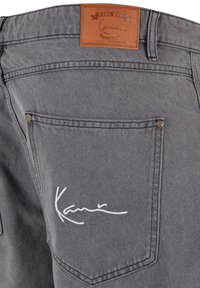 Pantaloni corti in denim grigio con una toppa in pelle con il marchio "Karl Kani". La tasca posteriore presenta dettagli ricamati bianchi e accenti metallici.