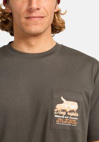 Camiseta de algodón gris con un bolsillo en el pecho que presenta un gráfico naranja de un coche y texto que detalla un servicio de surf. Con mangas cortas y cuello redondo.