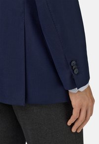 Námornícky textúrovaný blejzer s rozparkom, s štyrmi čiernymi gombíkmi na rukáve. Pod ním je viditeľný golier svetlomodrej pruhovanej košele.