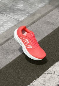 Bright coral New Balance running shoe with white sole on textured sidewalk near tactile paving and asphalt curb.