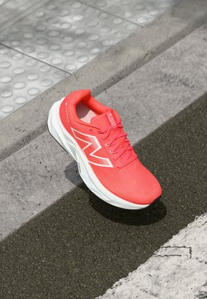 Svijetloružičasta New Balance trkačka cipela s bijelim potplatom na texturiranom pločniku u blizini taktilnog popločavanja i asfaltnog ruba.