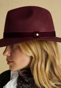 Pălărie din fetru vișinie cu bor larg, având o panglică din catifea și un accent metalic. Textură netedă, formă structurată.