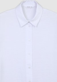 Camicia bianca a maniche corte con un tessuto a trama, colletto classico e chiusura frontale con bottoni bianchi. L'etichetta riporta "Terranova."