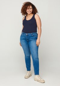 Top sin mangas de color azul marino oscuro combinado con pantalones ajustados de mezclilla azul claro. Los pantalones tienen bolsillos estándar y cierre de botón, usados con botas beige de cordones.