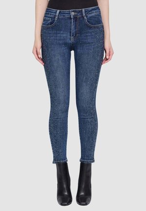 Donna con jeans aderenti blu scuro con dettagli in strass e stivaletti neri, in piedi davanti a uno sfondo semplice.