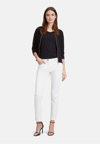 Giacca nera con zip, maglietta nera, pantaloni bianchi e tacchi a strappo neri. La modella sta in piedi contro uno sfondo bianco semplice.