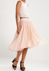 Beaded rosa Cropped Top mit hohem Kragen, kombiniert mit einem fließenden, geschichteten nudefarbenen Rock. Schwarze Knöchelriemen-Heels runden das Outfit ab.