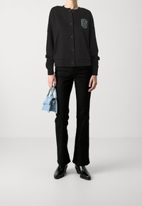 Zwarte cardigan met ronde hals, knopen aan de voorkant en een groen logo-patch. Gecombineerd met zwarte wijde jeans en een kleine blauwe handtas. Zwarte enkellaarsjes.