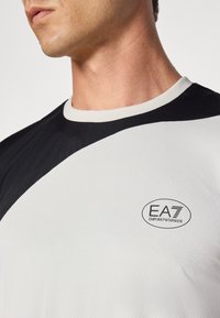 Trumpas rankovės sportinis marškinėliai, pasipuošę baltos ir juodos spalvų deriniu, tekstūruota medžiaga ir dideliu EA7 Emporio Armani logotipu krūtinėje.