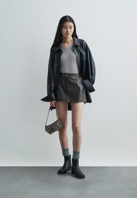 Camicia oversize in pelle nera, top grigio a coste, mini gonna marrone con cuciture, calzini grigi, stivaletti neri alla caviglia e una piccola borsa grigia.