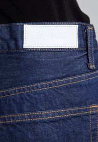 Tummansiniset farkut, joissa on erottuva keltainen ommeltu sauma ja valkoinen nahkalappu, jossa on kohokuvioituna "RE/DONE" takahihnassa.