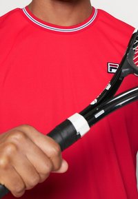 Henkilö, joka on pukeutunut punaisiin vaatteisiin, pitää mustaa, valkoisella kahvalla varustettua tennismailaa lähellä rintaa.