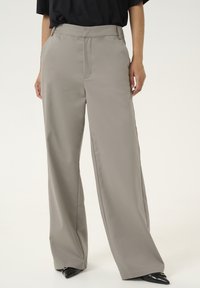 Un pantalon ample de couleur beige clair, fabriqué en tissu lisse. Il présente un pli central et des poches latérales, associé à des chaussures noires pointues.