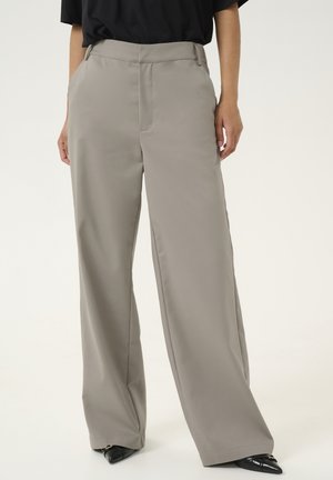 Un pantalon ample de couleur beige clair, fabriqué en tissu lisse. Il présente un pli central et des poches latérales, associé à des chaussures noires pointues.