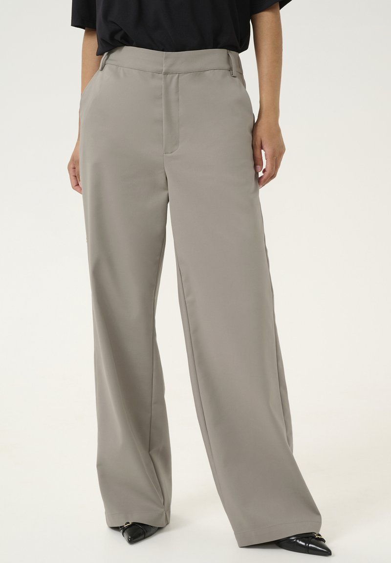 Un pantalon ample de couleur beige clair, fabriqué en tissu lisse. Il présente un pli central et des poches latérales, associé à des chaussures noires pointues.