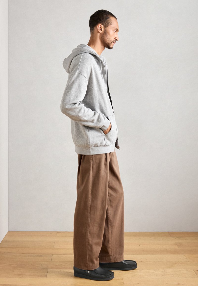 Grå zip-up hoodie med snodd i huvan, sidofickor, tillsammans med bruna vida byxor och svarta slip-on skor. Bakgrund av träparkett.