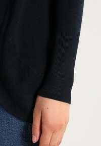 Dunkelblauer Strickpullover mit geripptem Ärmel, der eine Seitentasche und eine weiche Textur aufweist. Nahaufnahme, die eine Hand zeigt, die am Ärmel positioniert ist.