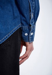 Camicia in denim blu con bottoni bianchi, cuciture visibili e polsini. Tessuto nero visibile sotto la camicia. Design pulito e casual.