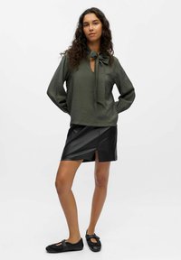 Olivengrüne, strukturierte Bluse mit einer Bindegarnitur am Ausschnitt, kombiniert mit einem schwarzen, synthetischen Lederrock mit einem Schlitz vorne und schwarzen Schuhen.