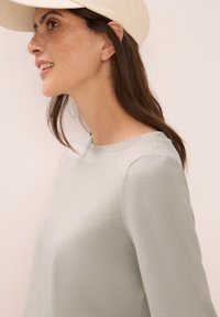 Light gray long-sleeve top with a subtle textured pattern, round neckline, and fitted sleeves, worn with a beige baseball cap.