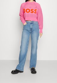 Růžový krátký svetr s designem síťoviny a oranžovým logem "BOSS", doplněný světle modrými širokými džínami a černými botami na platformě.