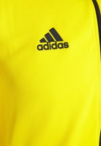 Geel polyester jack met een zwart geborduurd Adidas-logo, met een zachte textuur en een minimalistisch ontwerp. Afsluiting met rits.