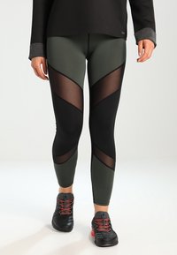 Legginsy w kolorze oliwkowym z wysokim stanem, z czarnymi prześwitującymi panelami i geometrycznymi wycięciami. Zestawione z czarnymi trampkami z czerwonymi akcentami.