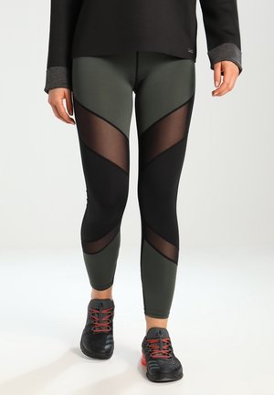 Leggings a vita alta di colore verde oliva con pannelli in rete nera trasparente e ritagli geometrici. Abbinati a sneakers nere con dettagli rossi.