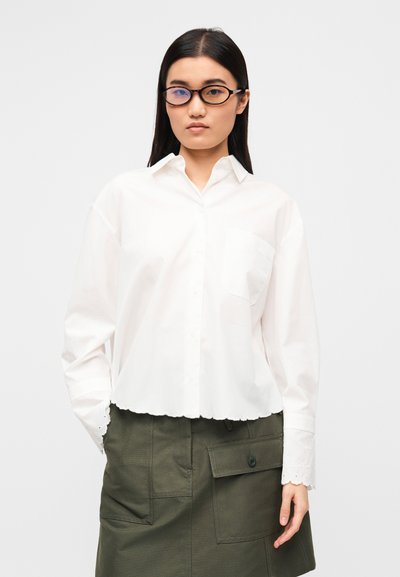 Λευκό κοντό πουκάμισο με κουμπιά, με κυματιστό τελείωμα και μία τσέπη στο στήθος, συνδυασμένο με φούστα ελιάς με μεγάλες τσέπες.