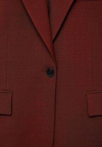 Close-up van een bordeauxrode blazer met één knoop, met gepunte revers en klepzakken, die de stoftextuur en stikdetails laat zien.