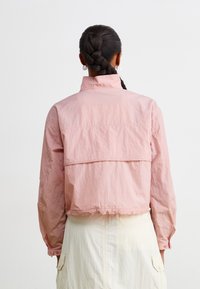 Lys rosa, kort jakke med høy krage, elastiske mansjetter og lagdelt bakdetalj. Laget av lett, teksturert stoff.