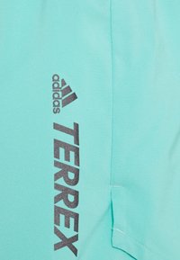 Logo de Adidas TERREX impreso en negro sobre tela turquesa clara con costuras visibles y borde de bolsillo.