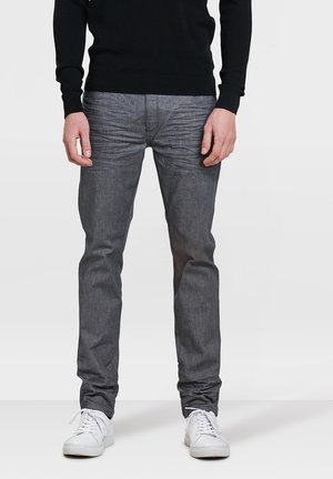 Mann trägt eng anliegende graue Jeans, weiße Sneakers und einen schwarzen Pullover mit langen Ärmeln, steht vor einem schlichten hellen Hintergrund.