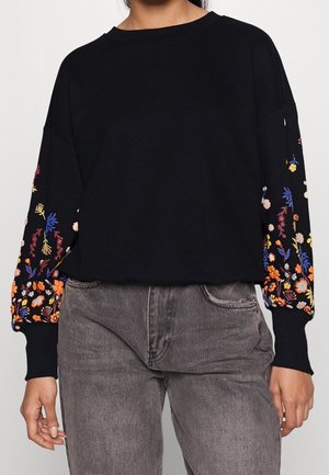 Zwarte trui met lange mouwen en kleurrijke bloemenborduursels op de mouwen, gedragen met grijze jeans met hoge taille.