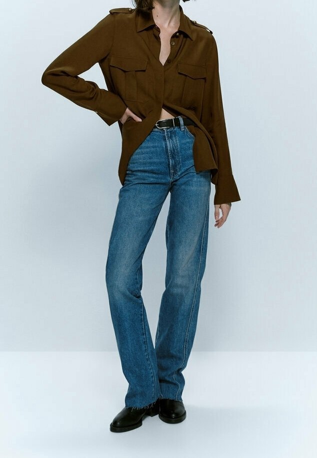 Femme portant une chemise marron à boutons, un jean bleu taille haute avec une ceinture noire et des chaussures noires, debout avec une main sur la hanche.
