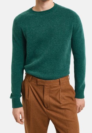 Zielony sweter z dzianiny z okrągłym dekoltem i prążkowanymi mankietami, w połączeniu z brązowymi spodniami w kant z teksturowanej tkaniny.