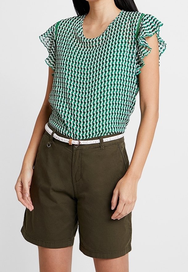 Femme portant un chemisier vert à motifs avec des manches à volants, un short vert olive et une ceinture blanche tressée, debout avec les bras détendus.