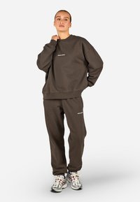 Brūns kokvilnas sporta krekls un atbilstošās sporta bikses ar brīvu piegriezumu. Balts logo uz krūšu un augšstilba. Modelis valkā metāliski sporta apavus.