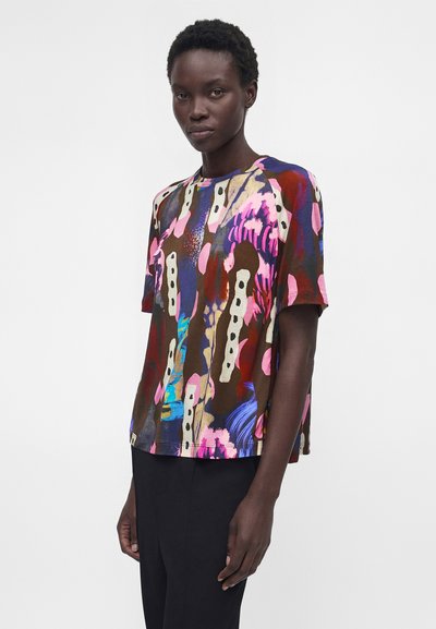Γυναίκα με κοντά κατσαρά μαλλιά που φοράει πολύχρωμο κοντομάνικο πουκάμισο με αφηρημένο σχέδιο και μαύρο παντελόνι, στέκεται μπροστά από απλό φόντο.