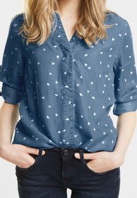 Denimskjorta i blått med vita fågeltryck, korta upprullade ärmar och krage. Mjuk textur och avslappnad passform.