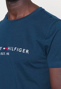 Närbild av en person som bär en blå Tommy Hilfiger-t-shirt med broderad logotyp och texten "EST. 1985" på bröstet.