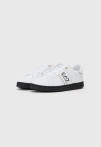 Sneakers bianchi con texture liscia, suola nera, dettagli oro e distintivo marchio EA7 sul lato. Design con lacci.