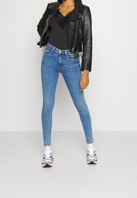 Schwarze Lederbikerjacke, schwarzes Oberteil mit Spitzenbesatz, blaue Skinny Jeans und weiße Sportschuhe mit roten und schwarzen Akzenten vor neutralem Hintergrund.