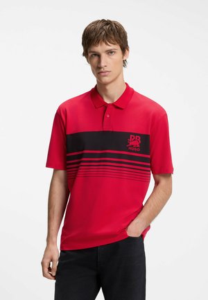 Giovane uomo che indossa una polo rossa con strisce orizzontali nere e un logo, in piedi con una mano in tasca su uno sfondo bianco.