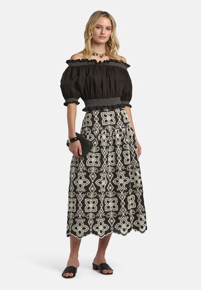 Blonde vrouw met een zwarte off-shoulder top met pofmouwen en een zwarte en witte midi-rok met bloemmotief, die een zwarte clutch vasthoudt.