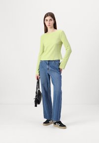 Lys grønn teksturert langermet topp kombinert med blå høyt liv rette jeans. Modellen holder en svart håndveske; iført svarte sko med hvite striper.