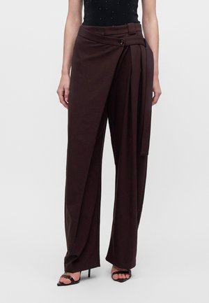 Femme portant un pantalon large marron foncé avec un cache-cœur asymétrique et un détail bouton à la taille, associé à des talons ouverts noirs.