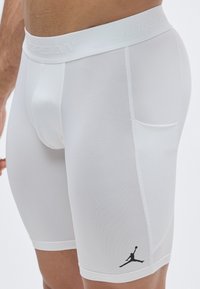 Λευκά αθλητικά σορτς με στενή εφαρμογή, κατασκευασμένα από ελαστικό ύφασμα, με ελαστική μέση που φέρει την ετικέτα "JORDAN" και μαύρο λογότυπο στην πλευρά.
